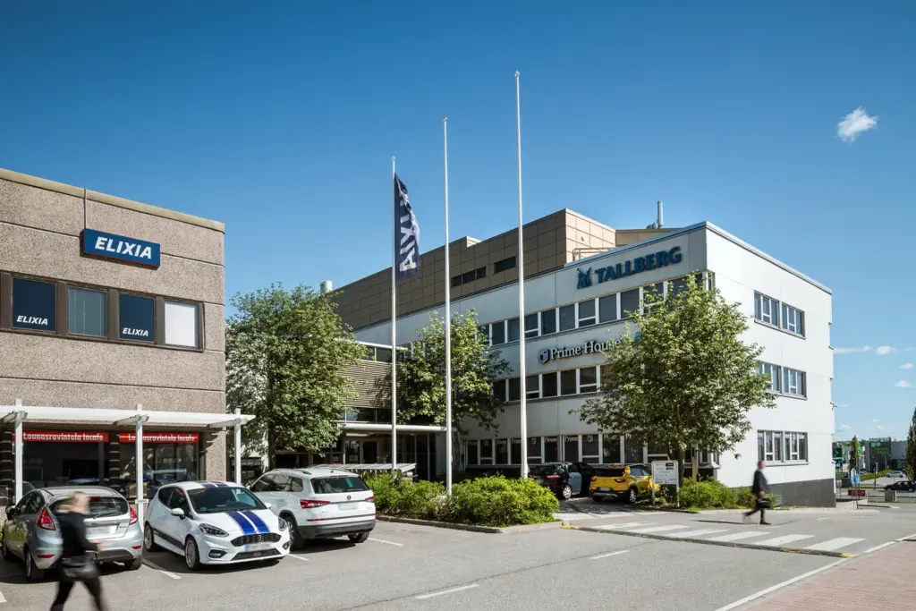 Matalan toimitilakiinteistön sisäänkäynti. Edessä on lipputankoja ja parkkipaikka.