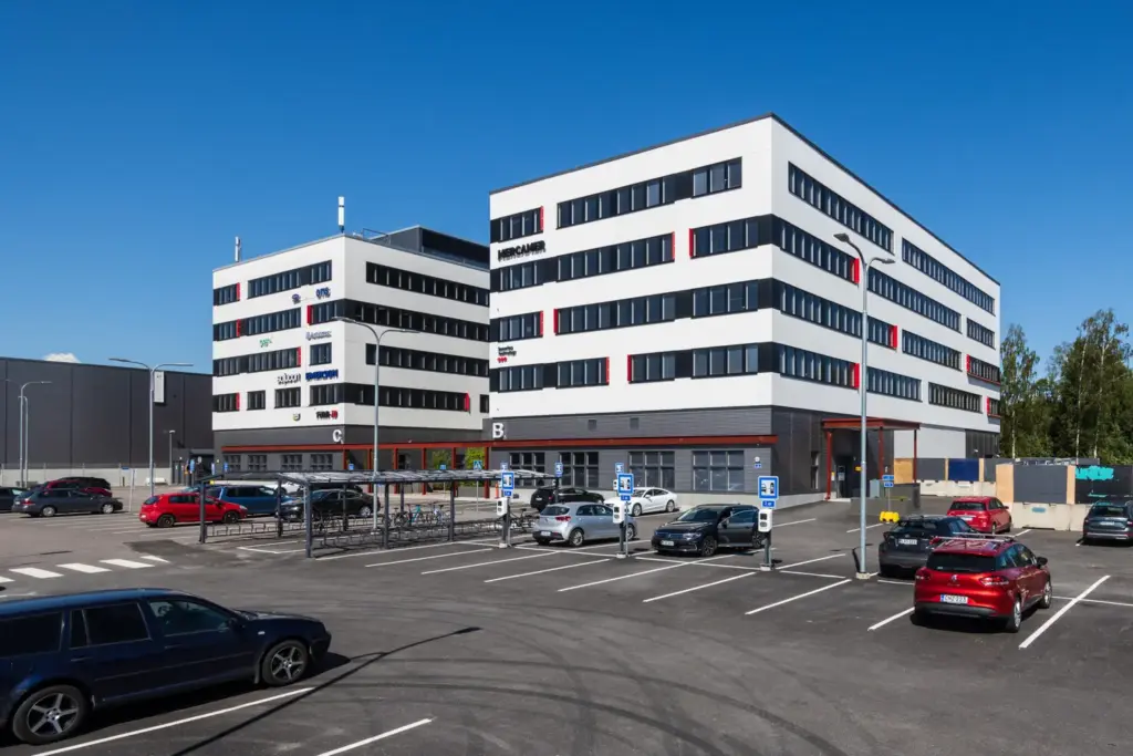 PRESSI Smart Premises C- ja B-talot pysäköintialueelta katsottuna. B-talon edessä on pyöräkatos ja pysäköintialueella sähköautojen latureita.