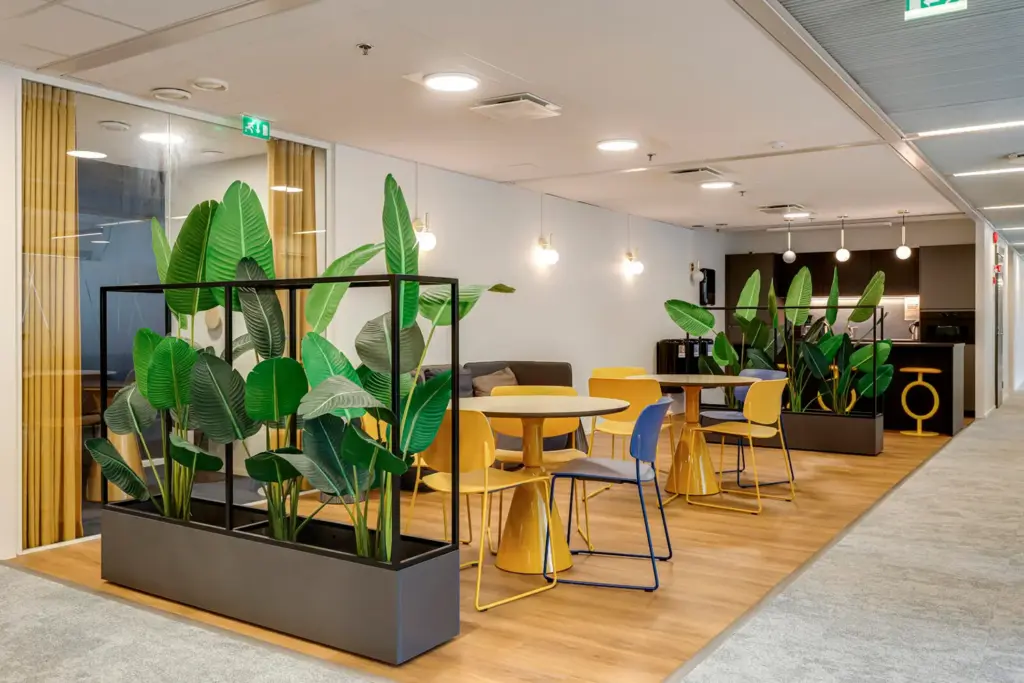 Viihtyisä taukotila Opus Business Parkin Bisneskylän Rantsu -pientoimistoalueella. Tilan keskellä kaksi pöytäryhmää ja molemmissa päissä viherkasvit. Taustalla baarikeittiö.